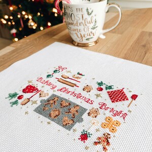 May include: White cross-stitch embroidery with a red and green Christmas theme. The embroidery depicts a variety of Christmas baking items, including gingerbread cookies, rolling pins, and a gingerbread house. The text "Baking Christmas Bright" is stitched in red.
