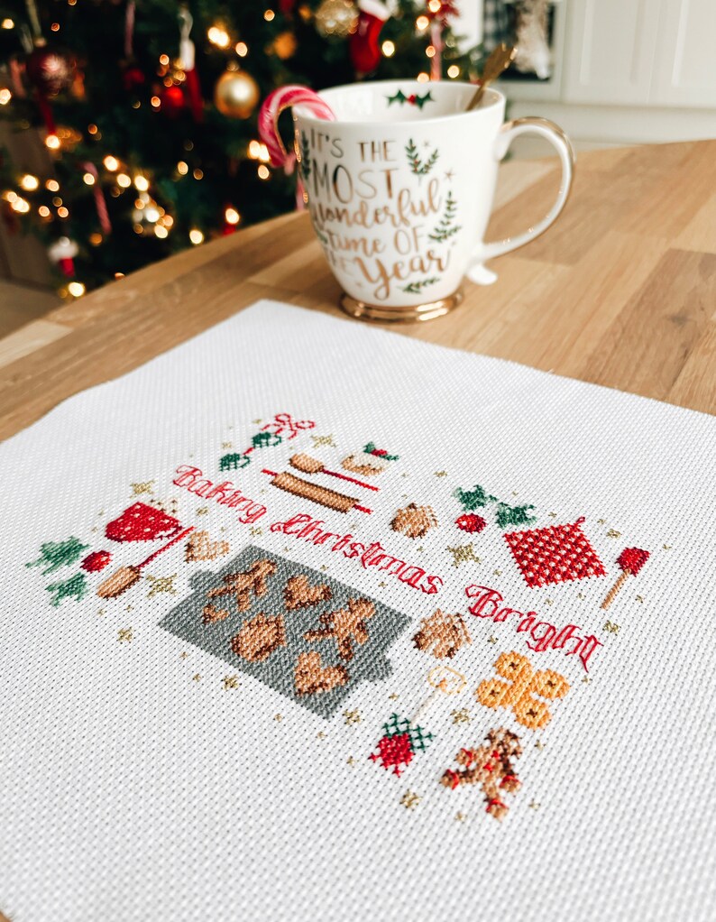 May include: White cross-stitch embroidery with a red and green Christmas theme. The embroidery depicts a variety of Christmas baking items, including gingerbread cookies, rolling pins, and a gingerbread house. The text "Baking Christmas Bright" is stitched in red.