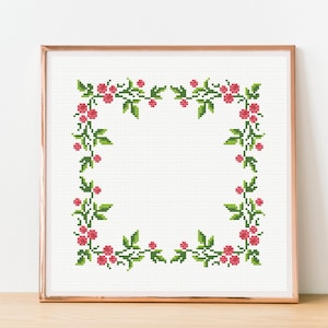 Könnte beinhalten: Ein Kreuzstichmuster mit einem floralen Rand aus rosa Blüten und grünen Blättern in einem weißen Rahmen.