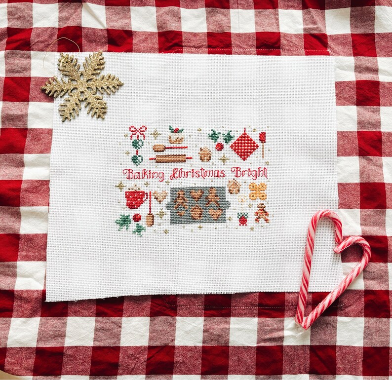 May include: White cross-stitch embroidery with a Christmas baking theme. The embroidery features a variety of festive baking items, including a gingerbread house, cookies, and a rolling pin. The text "Baking Christmas Bright" is stitched in red thread.