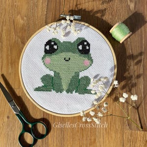 May include: A cross-stitch embroidery hoop with a green frog with big eyes and a pink blush. The frog is sitting on a white background.
