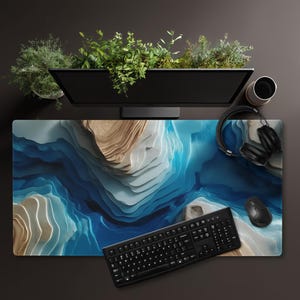 May include: A computer desk setup featuring a large mousepad with a blue and beige topographical design. A black keyboard, mouse, and headphones are on the pad. A computer monitor and a cup are visible, along with a planter with greenery.