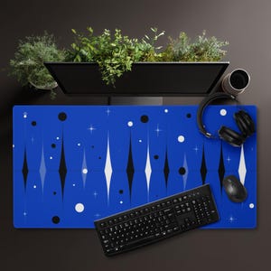Puede incluir: Una alfombrilla de escritorio azul con un patrón geométrico de formas de diamantes blancos y negros, círculos y estrellas. Un teclado, un ratón y unos auriculares negros están sobre la alfombrilla. Un monitor de ordenador y una planta son visibles.