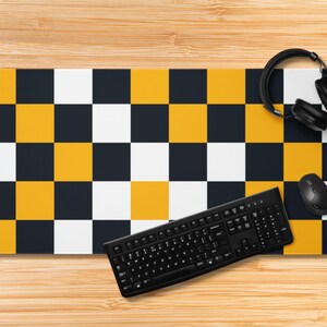 May include: A black keyboard, mouse, and headphones rest on a rectangular mousepad. The mousepad features a checkerboard pattern of yellow, white, and navy blue squares. The items are on a light brown wooden surface.
