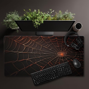 以下が含まれることがあります： 黒とオレンジ色のクモの巣デザインのマウスパッド、黒いキーボード、マウス、ヘッドフォン付き。コンピューターモニターとカップも机の上にあります。背景には緑の植物があります。