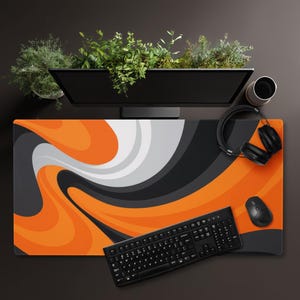 Op de afbeelding: Een rechthoekige bureaumat met een wervelend abstract patroon in levendig oranje, antracietgrijs, lichtgrijs en wit. De mat ligt onder een zwart computertoetsenbord, een zwarte draadloze muis en een zwarte hoofdtelefoon.