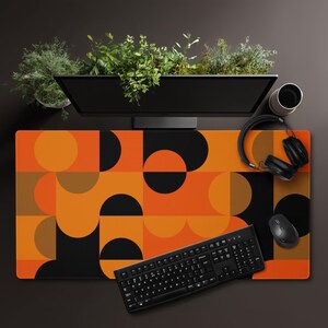 May include: A rectangular desk mat featuring a geometric pattern of overlapping circles and semicircles in shades of orange, brown, and black. The mat is positioned under a black computer keyboard, a wireless mouse, and a pair of black headphones.