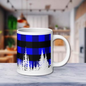 May include: White ceramic mug featuring a blue and black plaid pattern on the upper half. The bottom half of the mug displays a white silhouette of pine trees. The mug has a white handle and is set on a white marble surface.