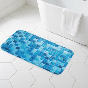 May include: A rectangular bath mat with a mosaic tile design in shades of blue. The mat is placed on a white hexagonal tile floor near a white bathtub and a window. A white towel hangs on the side of the tub.