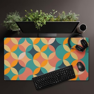 May include: A colorful desk mat with a geometric pattern of overlapping circles in teal, orange, pink, and yellow. A black keyboard, mouse, and headphones are on the mat. A computer monitor and a cup are visible.