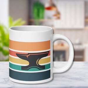 Könnte beinhalten: Weiße Keramiktasse mit einem Retro-Design, das eine schwarze Silhouette eines Ambosses auf einem Hintergrund aus orangefarbenen, gelben, grünen und blauen Streifen zeigt.