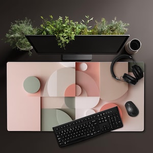 May include: A modern desk setup featuring a large, geometric patterned mousepad in shades of pink, beige, and green. A black keyboard, mouse, and headphones are on the pad. A monitor with greenery sits above the pad.