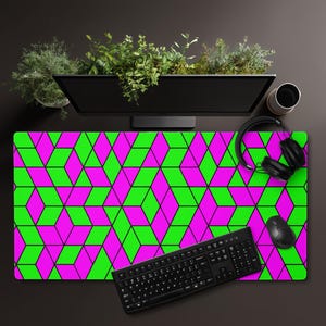 May include: A desk mat with a repeating pattern of green and magenta 3D cubes outlined in black. A black keyboard, mouse, and headphones are on the mat, with a computer monitor and a cup in the background.
