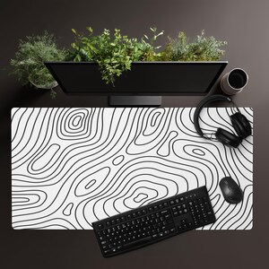 Puede incluir: Una alfombrilla de escritorio rectangular con superficie blanca y un patrón de líneas topográficas negras. La alfombrilla se sitúa bajo un teclado de ordenador negro, un ratón negro y auriculares negros sobre una superficie de escritorio marrón oscuro.