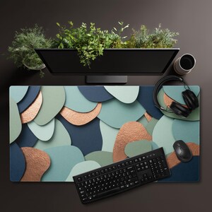 Pode incluir: Uma configuração de mesa com um mousepad grande e estampado com formas abstratas azuis, verdes e cor de cobre. Um teclado, um mouse e fones de ouvido pretos estão no mousepad. Um monitor de computador com vegetação em cima e uma xícara de café completam o espaço de trabalho.