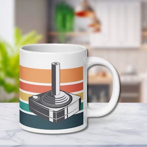 Pode incluir: Caneca de cerâmica branca com um design de joystick de videogame retrô. A caneca tem um fundo listrado em laranja, amarelo, verde e azul-petróleo. O joystick está em um esquema de cores cinza e preto.
