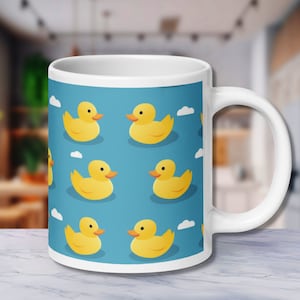 Könnte beinhalten: Weiße Keramiktasse mit blauem Hintergrund und einem sich wiederholenden Muster aus gelben Gummienenten.