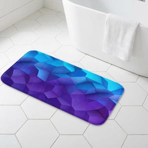 May include: A rectangular bath mat with a geometric design in shades of blue and purple. The mat is placed on white hexagonal tiles near a white bathtub. A white towel hangs on the side of the tub.