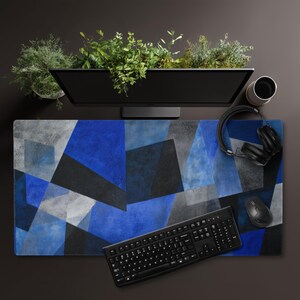 May include: A rectangular desk mat with a geometric design in shades of blue, gray, and black. A black keyboard, mouse, and headphones are on the mat. A computer monitor and a cup are also visible.