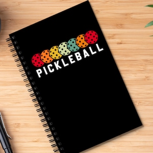 以下が含まれることがあります： 白文字で「PICKLEBALL」と書かれた黒いスパイラルバインダーノート。レトロなカラーリングで、カラフルなピクルボールが5つ並んでいます。