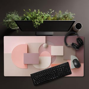 May include: A modern desk setup featuring a black computer monitor with greenery, a black keyboard, mouse, and headphones. The desk mat has a geometric design in shades of pink, beige, and brown. A coffee cup is also visible.