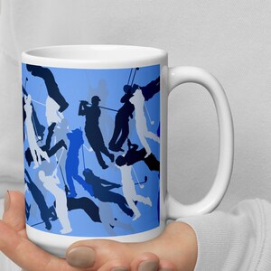 Puede incluir: Taza de café de cerámica blanca con fondo azul y un patrón de siluetas negras de golfistas jugando al golf.