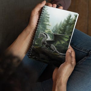 May include: A spiral bound notebook with a white cover featuring a detailed illustration of a silver dragon with green wings in a misty forest setting.
