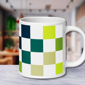 May include: White ceramic mug featuring a colorful checkerboard pattern in shades of green, blue, and cream. The mug has a curved handle and is set against a blurred background, suggesting a kitchen or dining setting.