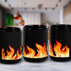 May include: Three black ceramic mugs featuring a flame design. The flames are orange, yellow and red. The mugs are on a white marble surface.