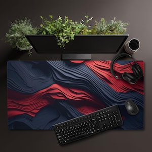 May include: A desk setup featuring a large, abstract patterned mousepad in shades of red and navy blue. A black keyboard, mouse, and headphones are on the pad. A computer monitor with greenery sits above.
