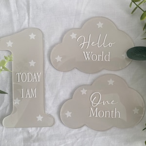 May include: Three grey acrylic signs with white stars and text. The first sign is a number one with the text "Today I am". The second sign is a cloud shape with the text "Hello World". The third sign is a cloud shape with the text "One Month".