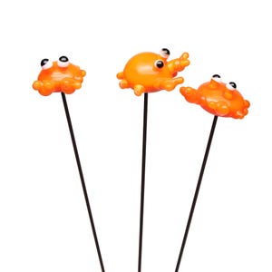May include: Three orange crab garden stakes with black stems and googly eyes. The crab stakes are made of a shiny plastic material and are set against a white background. The crab stakes are a fun and whimsical addition to any garden.