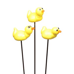 May include: Three yellow duck ornaments with orange beaks and black eyes, mounted on dark brown metal stakes. These ducklings are in a playful pose, ideal for garden or indoor decoration. The background is plain white.