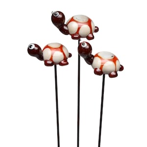 Könnte beinhalten: Drei dekorative Gartenstecker in Form von Schildkröten. Jede Schildkröte hat einen braunen Kopf und Beine, einen weißen Panzer mit roten Akzenten und schwarze Augen. Die Stecker sind dunkelbraun und scheinen aus Metall zu sein.