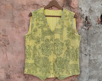 Puede incluir: Un chaleco sin mangas bordado en verde claro con un diseño de ciervos y figuras. El chaleco tiene un forro a rayas amarillas y blancas y está colgado de una percha de madera. El chaleco está abotonado y tiene dos bolsillos.