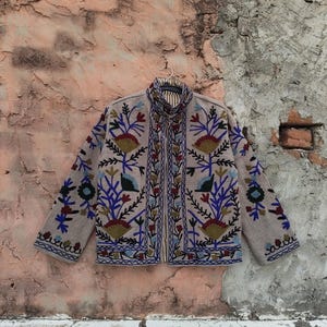 May include: Beige jacket with intricate floral embroidery in blue, red, and green. The jacket features a high collar and long sleeves, with detailed patterns along the front and cuffs. The jacket is hanging against a textured wall.