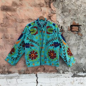 May include: Turquoise jacket with intricate floral embroidery in red, orange, and blue. The jacket features a mandarin collar and long sleeves. The embroidery creates a striking contrast against the turquoise fabric, showcasing a bohemian style.