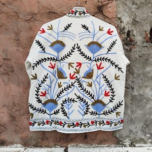 K&ouml;nnte beinhalten: Wei&szlig; bestickte Jacke mit floralem Design. Die Jacke hat blau, rot und schwarz gestickte Blumen und Bl&auml;tter. Die Jacke h&auml;ngt an einem Metallb&uuml;gel vor einer strukturierten Wand.