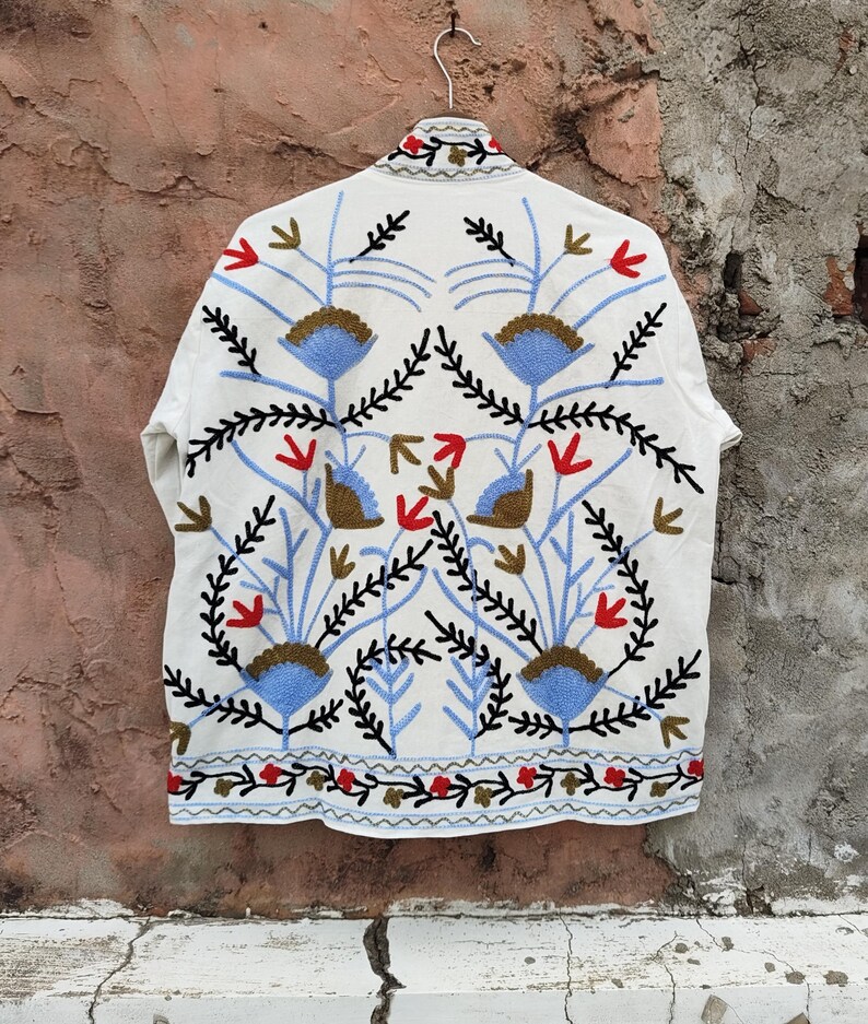 K&ouml;nnte beinhalten: Wei&szlig; bestickte Jacke mit floralem Design. Die Jacke hat blau, rot und schwarz gestickte Blumen und Bl&auml;tter. Die Jacke h&auml;ngt an einem Metallb&uuml;gel vor einer strukturierten Wand.
