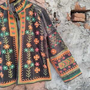 May include: A dark brown jacket with vibrant floral embroidery. The jacket showcases orange, green, and pink flower designs, with intricate patterns on the sleeves and trim. The jacket is suspended on a wooden hanger.