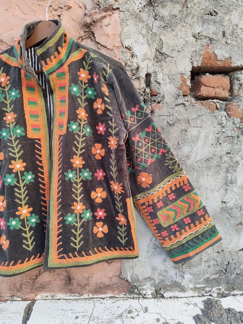 May include: A dark brown jacket with vibrant floral embroidery. The jacket showcases orange, green, and pink flower designs, with intricate patterns on the sleeves and trim. The jacket is suspended on a wooden hanger.