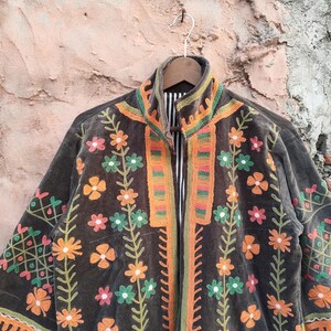 May include: A dark grey jacket with vibrant floral embroidery. The jacket showcases orange, green, and pink flowers, with matching trim on the collar and edges. The jacket is suspended from a wooden hanger.