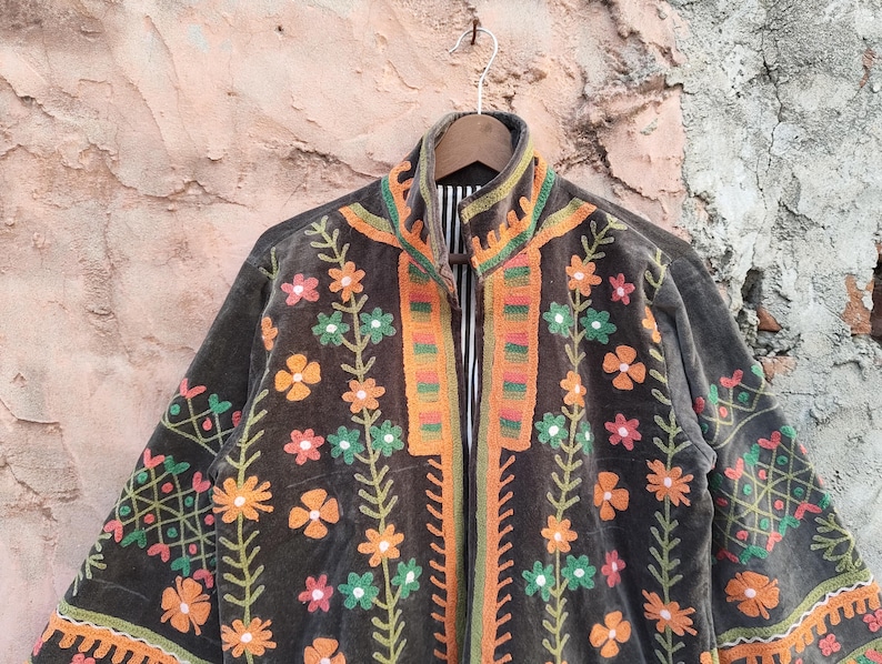 May include: A dark grey jacket with vibrant floral embroidery. The jacket showcases orange, green, and pink flowers, with matching trim on the collar and edges. The jacket is suspended from a wooden hanger.