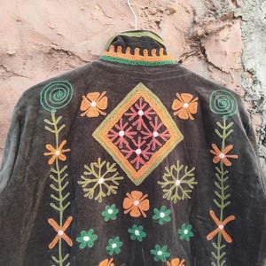 May include: A dark olive green jacket with detailed embroidery. The jacket showcases a central diamond design, framed by orange, green, and pink floral and geometric patterns. The collar and edges are also embellished with embroidery.