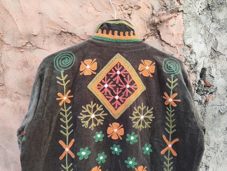 May include: A dark olive green jacket with detailed embroidery. The jacket showcases a central diamond design, framed by orange, green, and pink floral and geometric patterns. The collar and edges are also embellished with embroidery.