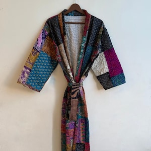 May include: A long-sleeved robe featuring a patchwork design made from various quilted fabric panels in shades of blue, purple, black, and gold. The garment includes a matching fabric tie belt at the waist and a light-colored interior lining.