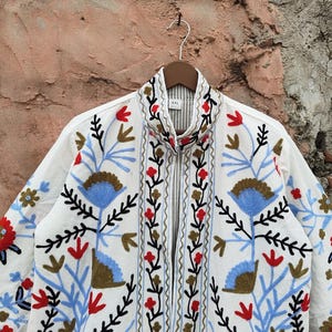 K&ouml;nnte beinhalten: Wei&szlig;e Jacke mit aufwendiger Blumenstickerei in Rot, Blau und Braun. Die Jacke hat einen Stehkragen, einen durchgehenden Rei&szlig;verschluss und h&auml;ngt an einem Holzb&uuml;gel. Die Stickerei umfasst stilisierte Blumen und Bl&auml;tter.