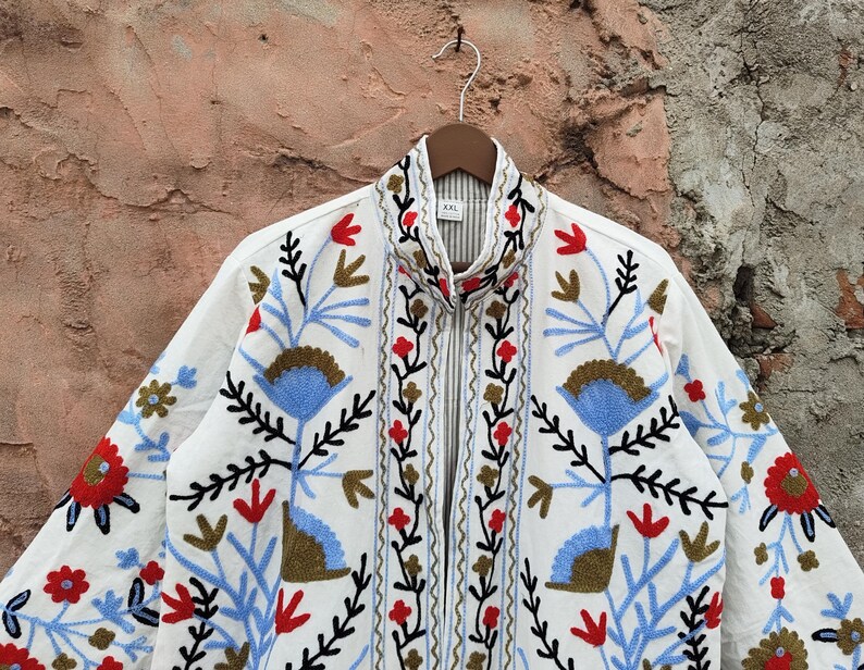 K&ouml;nnte beinhalten: Wei&szlig;e Jacke mit aufwendiger Blumenstickerei in Rot, Blau und Braun. Die Jacke hat einen Stehkragen, einen durchgehenden Rei&szlig;verschluss und h&auml;ngt an einem Holzb&uuml;gel. Die Stickerei umfasst stilisierte Blumen und Bl&auml;tter.