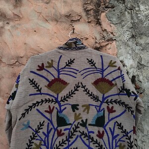 May include: A beige jacket with intricate floral embroidery in blue, red, green, and gold. The jacket is hanging on a silver hanger against a textured, weathered wall. The embroidery features stylized flowers and leaves.