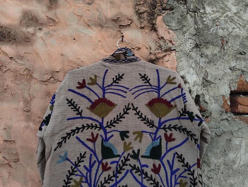 May include: A beige jacket with intricate floral embroidery in blue, red, green, and gold. The jacket is hanging on a silver hanger against a textured, weathered wall. The embroidery features stylized flowers and leaves.